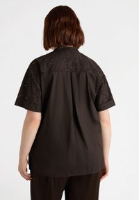 Camicetta marrone scuro con maniche corte, dettagli in ricamo a occhio sul retro e una vestibilità rilassata con orlo dritto.