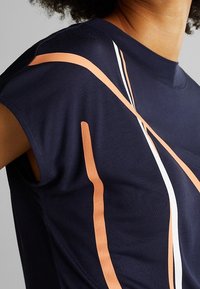 Marinblå tygtop med exponerade axelsömmar som har orange och vita abstrakta linjer som designaccent. Slät textur, avslappnad passform.