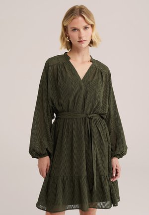 Femme aux cheveux blonds courts porte une robe longue manches, ceinturée, à motif vert foncé avec un décolleté en V sur un fond beige uni.