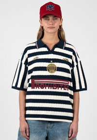 Mujer que lleva una camiseta tipo polo de rayas azul marino y blancas con el texto "PROHIBITED" y una gorra de béisbol roja con un logo azul, de pie frente a un fondo liso.