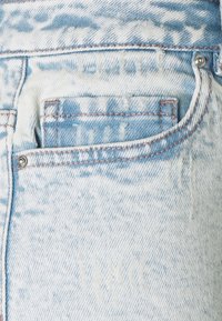 Světle modré oprané džíny mají náprsní kapsu s viditelným šitím, stříbrnými detaily a mírně vybledlou texturou.