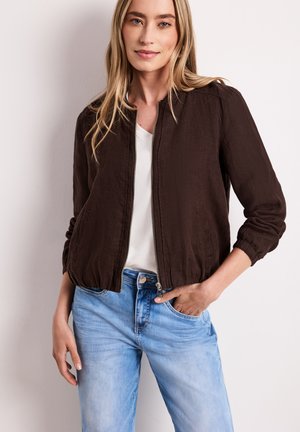 Ung kvinde med langt blond hår iført en mørkebrun lynlåstrøje over en hvid skjorte og lyseblå jeans, stående mod en hvid baggrund.