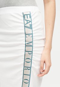 Λευκή φούστα με κάθετη μπεζ και πετρόλ ρίγα με λογότυπο "EA7 Emporio", φορεμένη με γκρι τοπ, εμφανίζοντας μέρος ενός χεριού που ακουμπά στο πλάι.