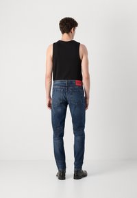 Tmavomodré denimové džínsy so slim fit, s červeným logom vzadu, doplnené čiernym tielkom bez rukávov a čiernymi topánkami.
