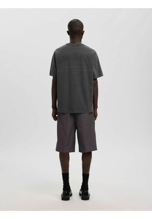 Personne debout, de dos à la caméra, vêtue d'un t-shirt oversize gris foncé et d'un short assorti longueur genoux sur un fond uni.