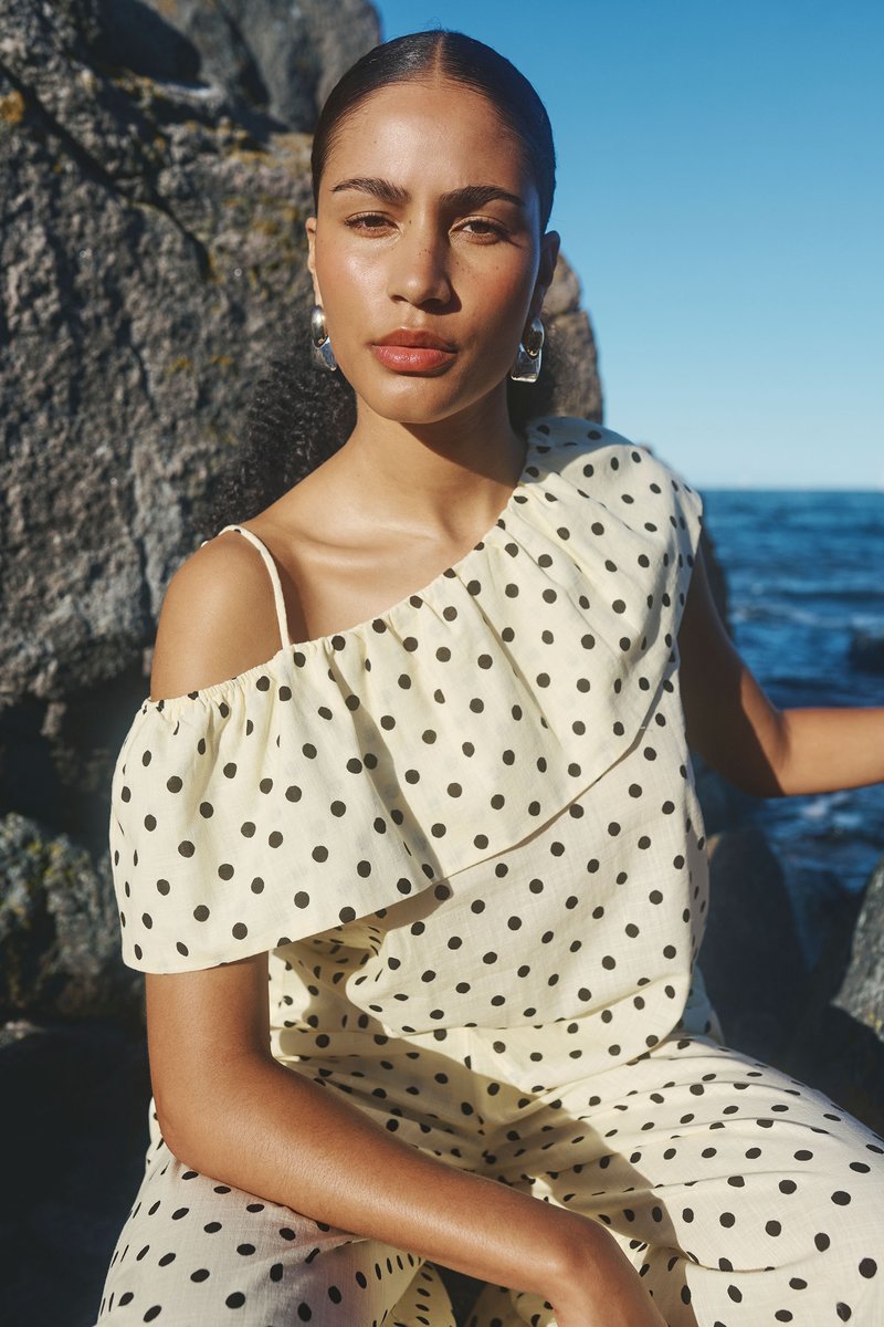 Femme aux cheveux lissés en arrière portant une robe crème à pois à une épaule et des boucles d’oreilles argentées en forme d’anneau, assise près d’un rivage rocheux et de l’océan sous un ciel clair.