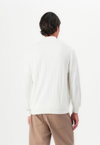 Man with short dark hair wearing a white ribbed sweater and light brown pants, viewed from behind against a plain white background.