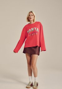 Červená mikina s nápisem "SANTA BABY" v barevných, texturovaných písmenech, doplněná hnědou koženou mini sukní a béžovými teniskami s červenými akcenty.