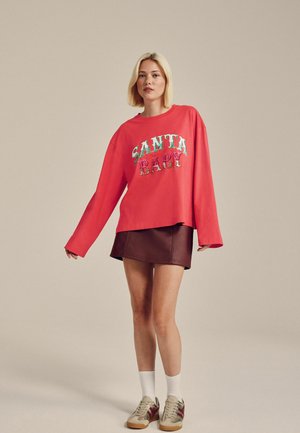 Roter Sweatshirt mit "SANTA BABY" in bunten, strukturierten Buchstaben, kombiniert mit einem braunen Lederminirock und beigen Sneakers mit roten Akzenten.