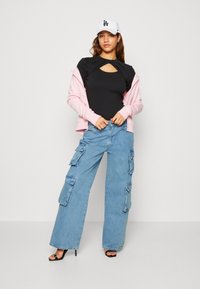 Top nero a ritaglio, felpa rosa con zip, pantaloni cargo azzurri chiaro con grandi tasche e tacchi a strappy neri; indossa un cappellino bianco con logo.