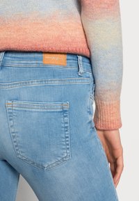 Jeans de color azul claro con una etiqueta de cuero, diseño ajustado y bolsillos traseros; combinados con un suéter de punto en degradado de rosa a azul.