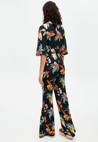 Blomstrede jumpsuit med brede ærmer, sort baggrund, farverigt blomsterprint i røde, gule og grønne nuancer, løs pasform og flagrende bukser.
