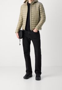  gepolsterte olivgrüne Jacke mit Kapuze, kombiniert mit schwarzen Jeans und schwarzen Sneakern. Zubehör: kleine schwarze Umhängetasche.