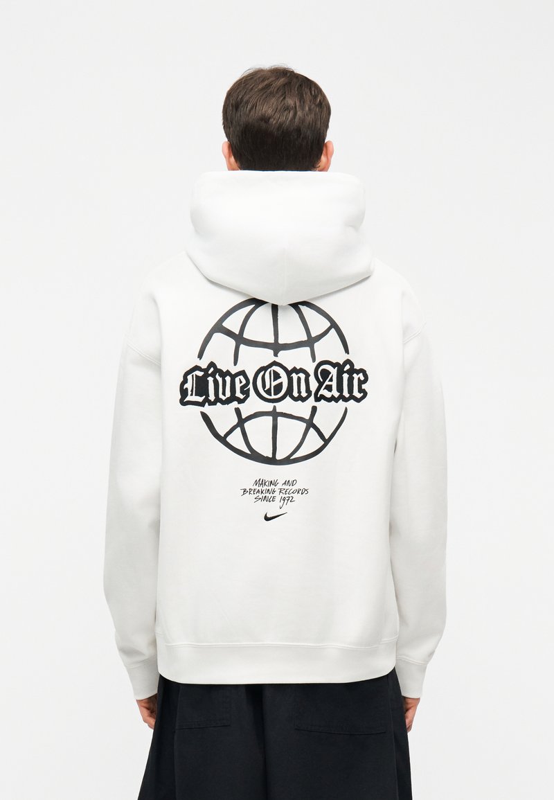 Person trägt weißen Kapuzenpullover mit schwarzem Globus-Grafik und der Aufschrift "Live On Air" auf dem Rücken, kombiniert mit schwarzer Hose, Rücken zugewandt.
