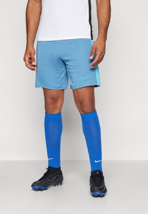 Completo sportivo da uomo con pantaloni corti azzurri chiari caratterizzati da un'accenno blu scuro e logo, abbinati a Calze alte blu e scarpini da calcio neri.