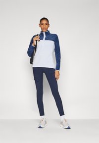 Fleece-pullover i marineblå og hvit med høy krage, sammen med marineblå cargo leggings og lyse sneakers med akenter. Modell holder svart veske.