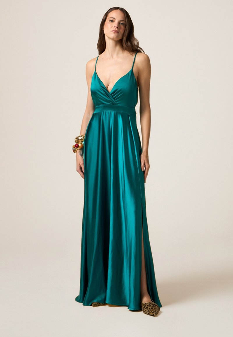 Abito di raso teal con spalline sottili, profondo scollo a V e vita arricciata. Due spacchi laterali e silhouette fluida. Accentuato con bracciali audaci.