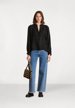 Femme debout portant un chemisier noir à manches longues, un jean droit bleu, des chaussures noires, tenant un sac à main tissé à motif chevron.