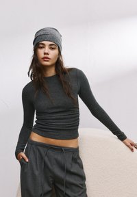 Graues Langarmoberteil mit Raffungen und Rundhalsausschnitt, kombiniert mit lockeren grauen Hosen. Eine graue Mütze vervollständigt den Look.