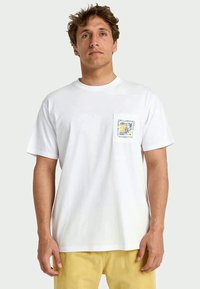 Man met tousled haar draagt een witte T-shirt met een klein kleurig Billabong-logo op de borstzak en gele broek.