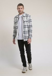 Chemise à carreaux noir et blanc avec des boutons, superposée sur un T-shirt blanc. Associée avec un jean noir et des baskets grises.