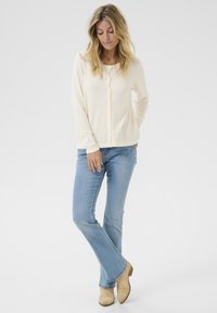Cardigan crema con bottoni e maniche lunghe, abbinato a jeans a zampa d'elefante blu chiaro e stivali alla caviglia beige. Design semplice e texture morbida.