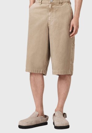 Beige bomuldsshorts med afslappet pasform, knælange, med sidelommer og synlige syninger. Kombineret med lyse træsko.