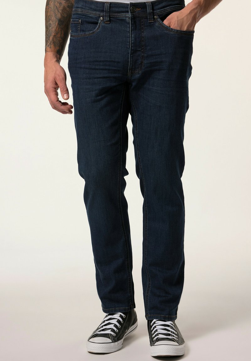 Jeans in denim blu scuro con vestibilità slim, cinque tasche, cuciture chiare e chiusura classica con bottone e zip, abbinati a sneakers nere.