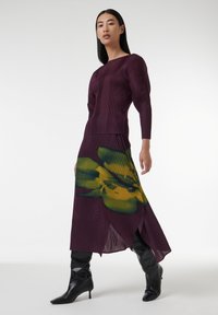 Largo vestido plisado en púrpura profundo con un estampado floral verde en la falda. Presenta un cuerpo ajustado y un dobladillo dividido, acompañado de botas negras hasta la rodilla.