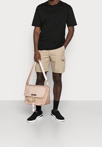Personne portant un t-shirt noir, un short cargo beige, des chaussettes noires et des baskets noires, tenant un sac en bandoulière beige par une sangle sur le côté.