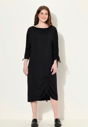 Robe midi noire avec un col rond, des manches trois-quarts, des détails froncés sur les côtés avec des liens, et un ourlet froncé. Texture de tissu lisse.