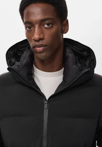 Giacca a vento nera con zip frontale e cappuccio con coulisse, caratterizzata da una texture liscia e un design leggero, indossata sopra una camicia bianca.