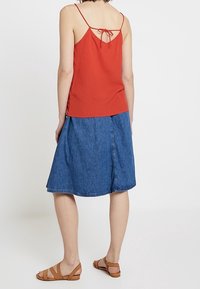 Femme portant un haut sans manches rouge avec un nœud dans le dos, une jupe en denim bleu à mi-cuisse et des sandales plates marron, se tenant de dos.