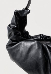 Fekete bőr táska puha, gyűrött textúrával, ívelt füllel és az egyik oldalon látható zipzárral.