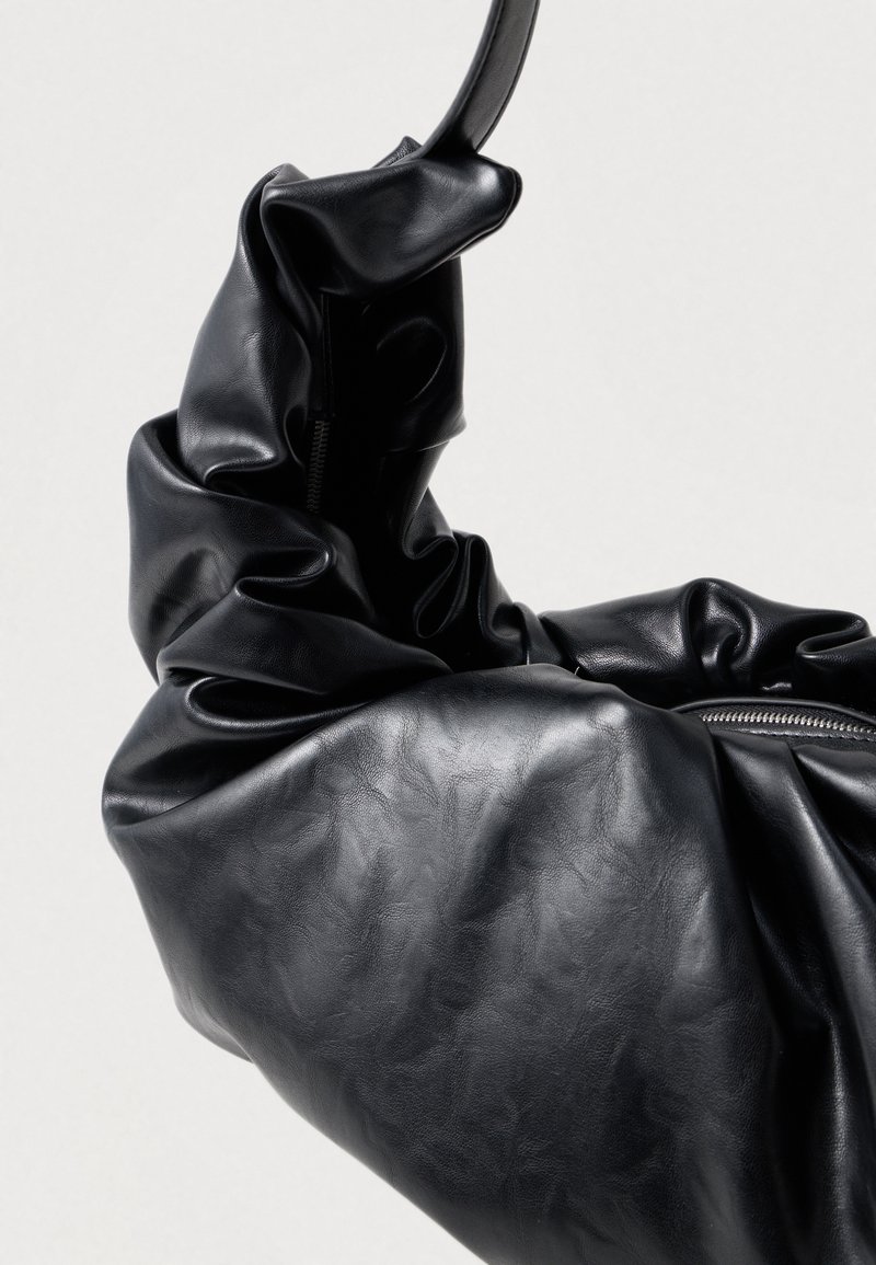 Schwarze Lederhandtasche mit einer weichen, zerknitterten Textur, die einen geschwungenen Griff und ein sichtbares Reißverschlusdetail auf einer Seite aufweist.