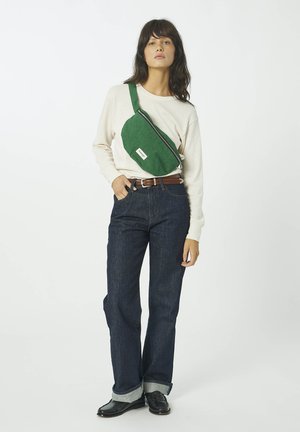 Femme portant une chemise crème à manches longues, un jean bleu foncé retroussé, des chaussures noires, une ceinture marron et un sac bandoulière vert sur un fond blanc.