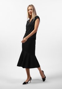 Čierny priliehavý top s detailom na zips, doplnený čiernou rybou sukňou s rozšíreným lemom. Vysoké čierne topánky na podpätku dokončujú outfit.
