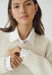 Cremefarvet ribstrikket sweater lagdelt over en hvid stribet skjorte med krave og juvelbesatte manchetter. Blød tekstur og skræddersyede design detaljer.