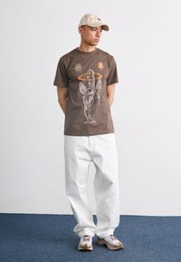 Brun bomulds T-shirt med hvid grafisk print og orange accenter. Sammen med løstsiddende hvide bukser og grå sneakers.