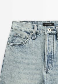 Jean en denim bleu clair avec fermeture à boutons sur le devant, passants pour ceinture, petite poche à monnaie, et une étiquette Massimo Dutti à l'intérieur de la ceinture.