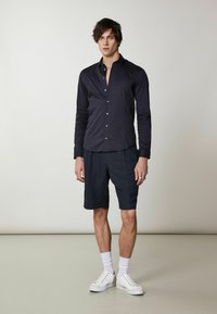 Maglietta a maniche lunghe blu con bottoni, abbinata a pantaloni corti scuri. Il modello indossa scarpe da ginnastica bianche con calzini alla caviglia. Design semplice, texture liscia.