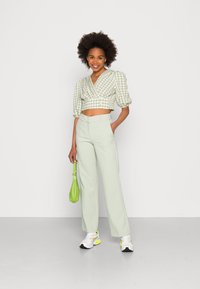 Blusa curta xadrez verde com mangas bufantes, combinada com calças largas verde claro. Segurando uma pequena mala verde e usando tênis.