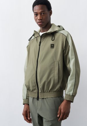 Άνδρας που φοράει ανοιχτό πράσινο μπουφάν με φερμουάρ και κουκούλα, στέκεται μπροστά σε λευκό απλό φόντο.