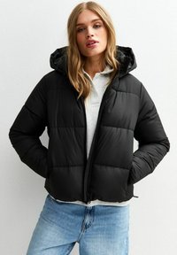 Zwarte pufferjack met een capuchon, met een gequilt ontwerp en een rits aan de voorkant. Gecombineerd met een lichte top en blauwe jeans. Gladde textuur.