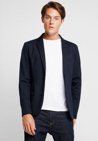 Blazer navy con un diseño a medida, dos botones frontales y un único bolsillo en el pecho. Llevado sobre una camiseta blanca, combinado con jeans oscuros.