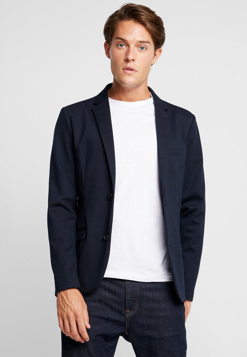 Blazer navy con un diseño a medida, dos botones frontales y un único bolsillo en el pecho. Llevado sobre una camiseta blanca, combinado con jeans oscuros.