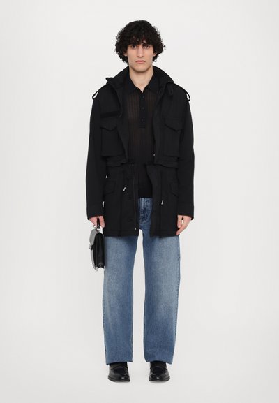 Zwarte, gevoerde jas met meerdere voorzakken, gecombineerd met een zwart shirt en lichtblauwe wijde jeans. Zwarte schoenen en een kleine tas.