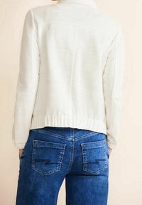 Biała teksturowana kurtka z kołnierzem, prążkowanym dołem i długimi rękawami, w zestawieniu z niebieskimi dżinsami z ukośnymi tylnymi kieszeniami.