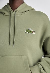 Sudadera verde oliva con cordones y un bolsillo canguro. Presenta un logo bordado de cocodrilo verde en el pecho. Textura de tela suave.