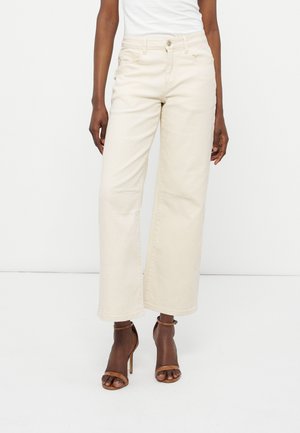 Γυναίκα που φοράει κρεμ παντελόνι με ψηλή μέση και φαρδιά πόδια και καφέ σανδάλια με ψηλό τακούνι, στέκεται μπροστά σε λευκό απλό φόντο.